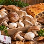 Variedades de setas comestibles frescas, como champiñones y setas de ostra, sobre una mesa rústica de madera, con una empanada gallega de setas al fondo.